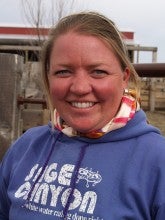 Cathy stands outside at her ranch in front of a fence with metal bars. She wears a blue hoodie that says "sage canyon" and a red and white striped scarf.