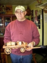 Frank Bettencourt stands and holds a small wooden vehicle replica. He wears a tan baseball cap, red sweater, and jeans.