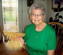 Alene Rucker sits at a dining room table and holds an example of her decorative wheat weaving. She is wearing a green t-shirt.