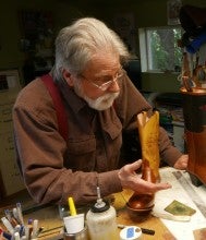 D.W. Frommer II sits at his workbench and gestures at a pair of brown cowboy boots. He wears a brown collared shirt with brown suspenders.