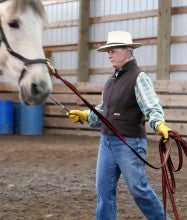 Alice Trindle leads a white horse in an indoor arena. She is wearing a white hat, a brown vest, blue jeans, and cowboy boots.