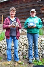 Cindy Kopcinski and Janis Smith stand outside in front of a dark brown building and a pile of thunder eggs and hold large thunder egg slices