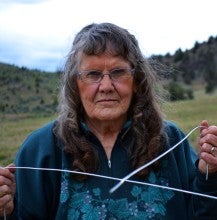 Betty Woodward stands outside and holds two water witching rods in Mitchell, Oregon. She wears a green quarter zip jacket with leaves on it.