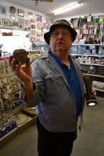 Dan Cannon stands inside his tire shop, Cannon's Tire Shop, holding a thunder egg in his right hand and a flashlight in his left. He wears a blue collared shirt over a blue polo shirt and a green fishing hat.