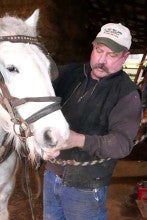 Duane Van Dyke bridles a white horse. He wears blue jeans, a black vest over a black long sleeved shirt, and a white and green baseball cap.