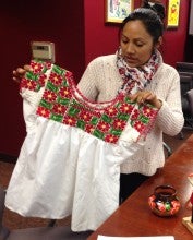Dorotea Lopez stands and holds a red, green, and white embroidered blouse. She wears a cream sweater and a patterned scarf.