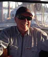 Dennis Best sits smiling in a boat with the windshield in the background. He wears a striped white jacket, sunglasses, and a baseball cap.