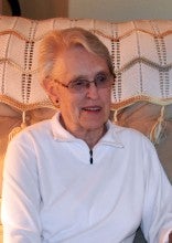 Alice Smith sits on a beige armchair in her home in Vernonia, Oregon. She wears a white fleece collared sweatshirt.