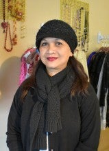 Chan-Phirath Farley stands in front of traditional Cambodian wedding attire at Fina Salon in Beaverton, Oregon. She wears a black jacket, black knitted scarf, and black knitted hat.