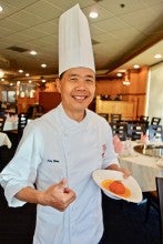 Andy Wong stands in his Portland restaurant and poses with an orange fruit dish with sauce. He wears a white chef's uniform.