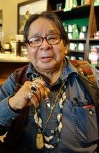 Ed Edmo sits in front of green metal shelves. He wears a dark blue long sleeve shirt, a patterned vest, two shell necklaces, a medicine bag, and glasses.