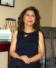 Azar Salehi sits in a room with chairs and a table in the background with her hands clasped. She wears a black dress, black tights, and a yellow pendant.