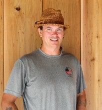 Brian Krehbiel stands and poses against a wood wall. He wears a gray shirt and a brown straw hat.