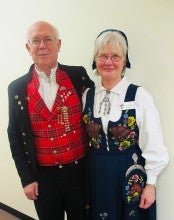 David and Claire stand against a white wall wearing traditional Norwegian folk outfits. David wears black pants, a red plaid shirt, white undershirt, and black jacket. Claire wears an embroidered navy blue dress over a long sleeve white shirt.