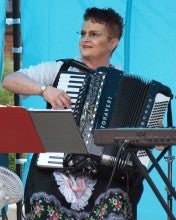 Joan Blaser plays an accordion in front of a blue backdrop, a music stand, and a keyboard. She wears a traditional black and white lace floral dress.