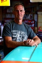 Brad Finley stands in his workshop with his forearm resting on a blue surfboard. He wears a dark gray t-shirt with a white surfing skeleton wearing a sombrero.