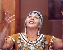 Esther charismatically tells a story, gesturing upwards with both of her hands. She wears a traditional leather outfit made with shells and beads as well as a beaded cap.