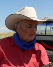 Barbara Nelson sits in the back of a truck playing a guitar. She wears a red shirt, blue bandana, and white cowboy hat.