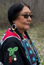 Roberta Kirk stands outside on grass. She is wearing black regalia with red, green, and blue beaded adornments as well as hoop earrings and sunglasses.