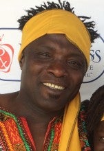 Alseny Yansane stands against a white background with a logo. He wears a yellow headband and multicolored, patterned clothes. 