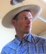Jack Armstrong wears a plaid blue long sleeve shirt and a white cowboy hat.