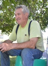 Gregorio Cortaberria sits outside on a teal bucket and holds a mason jar. He wears a yellow t-shirt with suspenders and blue jeans.