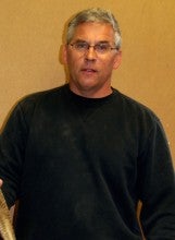Joe Dabulskis stands indoors against a light brown wall. He wears a black crewneck sweater.