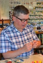 Jeff Perin stands in his shop in Sisters, Oregon, and ties a fly. He wears a blue and white plaid collared shirt.