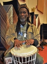 Yingwana Khosa demonstrates South African drumming. He is wearing an olive green outfit shirt and pants and a black beanie.