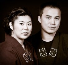 Michelle Fujii and Toru Watanabe pose against a black background. They both wear black robes with kanji characters on them.