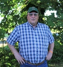 Jake Fallesen stands outside in front of a tree and wears a blue and white plaid shirt and green baseball cap.