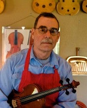Jeff Manthos stands in his workshop holding a violin. He wears a blue collared shirt and a red apron.