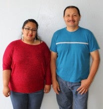 A woman, wearing a red shirt, and a man, wearing a blue shirt, stand in front of a white wall.