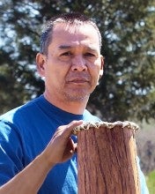Dean Adams kneels outside with a juniper bark basket resting on his knee, and two more of the same baskets on his right and left. He wears a blue shirt and blue jeans.