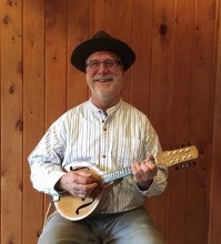 Bryan Ibach sits in front of a wood wall while holding a tan mandolin instrument and wearing a black hat, white striped shirt, and gray pants.