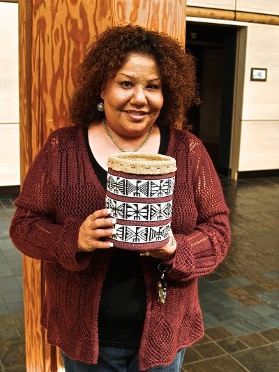 Natalie Kirk stands in front of a wooden pole wearing a black shirt and red cardigan. She holds a small white, tan, and black woven basket.