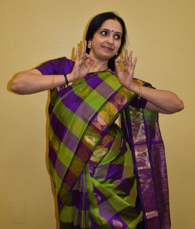 Anuradha Ganesh stands and poses in an expressive dance gesture against a tan wall. She wears a purple and green checkered sari.