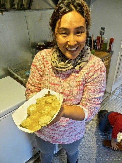 Ang Diki Sherpa stands in a kitchen and presents a paper tray of Nepalese momo dumplings covered with a yellow sauce. She wears a pink sweater, gray leggings, and a gray scarf with black cats on it.