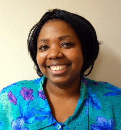 Anne-Marie Urukundo stands and poses against a white wall. She wears a blue floral collared shirt.