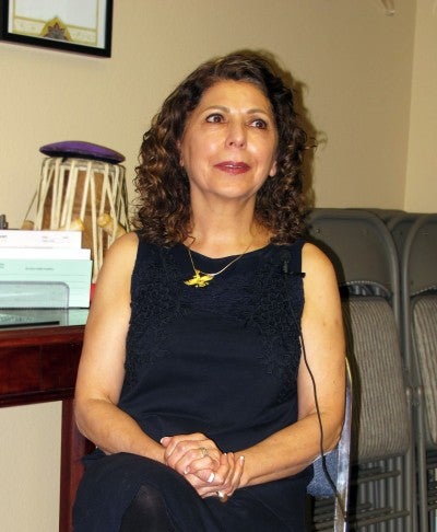 Azar Salehi sits in a room with chairs and a table in the background with her hands clasped. She wears a black dress, black tights, and a yellow pendant.