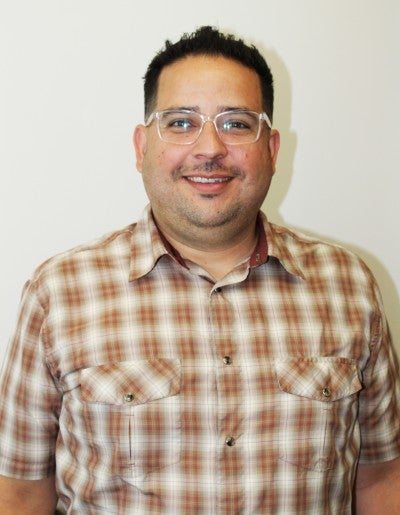 Arturo Zavala stands and poses against a white wall. He wears a brown and white plaid button-up shirt.