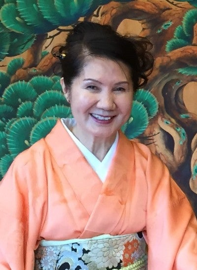 Masumi Timson sits behind a large wooden string instrument and in front of a wooden wall with painted tree imagery. She wears a peach kimono with a white floral obi.