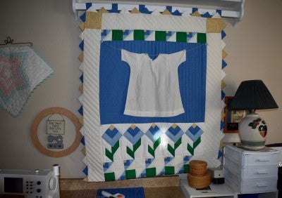 A hanging quilt with a white background, a large blue square in the middle containing a white shirt, and blue, yellow, and green flowers at the bottom.