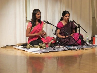 Anita sits in front of a microphone on a dance floor with another girl, both wearing pink traditional outfits.