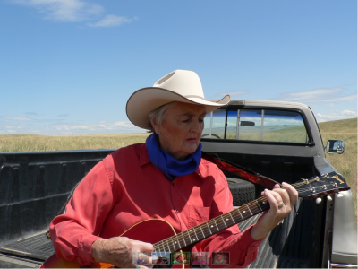 Barbara sits in the back of a truck playing a guitar and wearing a red shirt, blue bandana, and white cowboy hat.