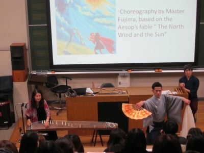 A woman plays a large wooden string instrument and a man dances with an orange hand fan.