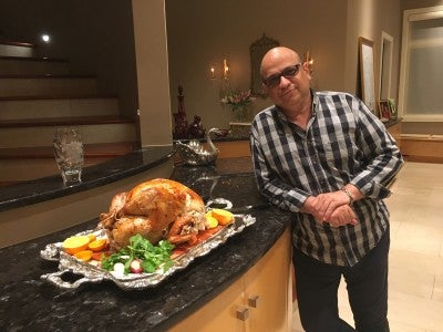 Ali, wearing a plaid button-down shirt, stands in a kitchen with a cooked turkey on the counter next to him.