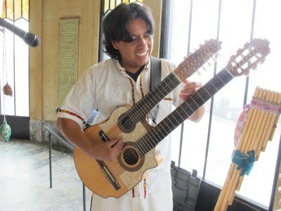  Alex Llumiquinga Perez holds a double necked guitar with a microphone and wooden pipes in the background.