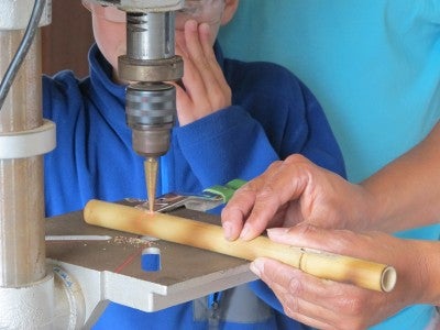 Person making a wooden pipe at a machine. A child watches wearing goggles and a blue fleece sweater.