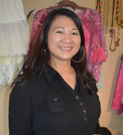 Amara Sim headshot wearing a black blouse with dresses and necklaces in the background